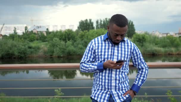 Чернокожий мужчина смотрит в камеру телефона и улыбается на набережной в парке . — стоковое видео