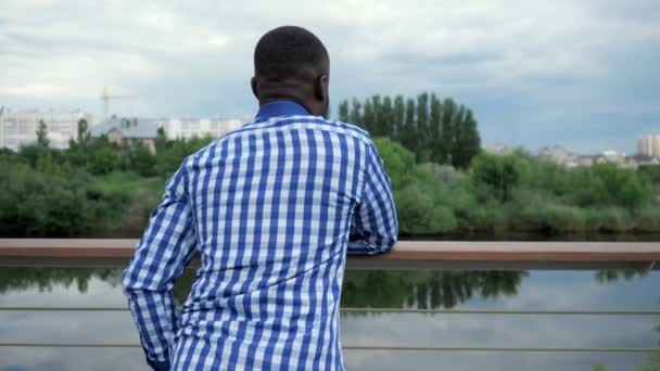 Black man staat op de stad waterkant nabij hek in Park en bewondert uitzicht op de stad. — Stockvideo