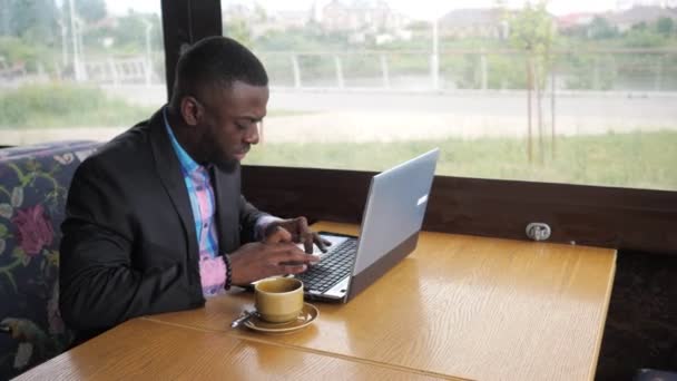 Black businessman is working typing a message on laptop sitting in summer cafe. — Stock Video