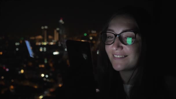 Mujer sonriente está mirando el teléfono inteligente cerca de la ventana con vista a la ciudad, vista lateral . — Vídeos de Stock