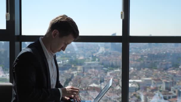 Ein fiktiver Arbeiter im Anzug tippt auf seinem Laptop am Fenster mit Blick auf die Stadt. — Stockvideo