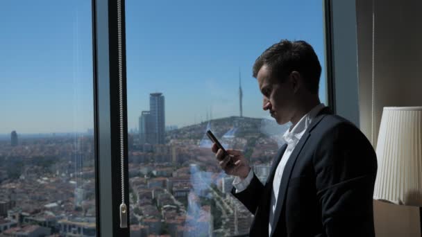 Joven empresario en traje de oficina está buscando teléfono cerca de la ventana con vista panorámica de la ciudad . — Vídeo de stock