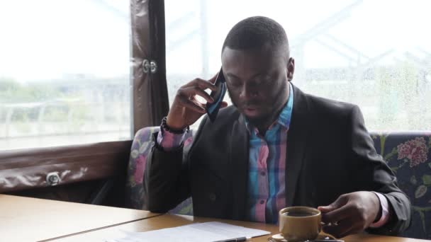 Afro american businessman is calling on smartphone and drinking coffee in cafe. — Stock Video