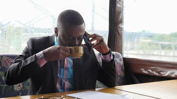 Afro americano uomo d'affari sta chiamando su smartphone e bere caffè in caffè . — Foto Stock
