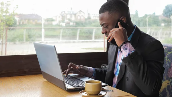 L'uomo d'affari nero lavora su computer portatile e telefona smartphone si siede in caffè con caffè . — Foto Stock