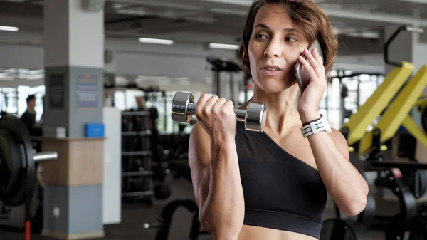 Ώριμη γυναίκα κάνει άσκηση δικέφαλων με βαράκια στο γυμναστήριο και μιλώντας τηλέφωνο. — Φωτογραφία Αρχείου
