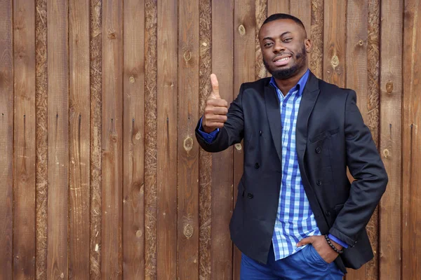 Black afro american smiling man shows gesture thumb up on dark wood background.