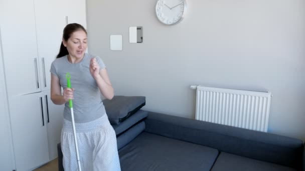 Mutlu kadın paspas kullanarak zemin yıkıyor, bir şarkı söylüyor ve modern dairesinde dans ediyor.. — Stok video