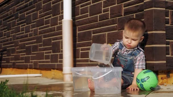 Niño con pelo corto y claro vierte agua de cubo de plástico — Vídeos de Stock