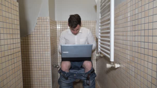 Молодой человек трудоголик работает в туалете на компьютере, печатая что-то и думая: . — стоковое видео