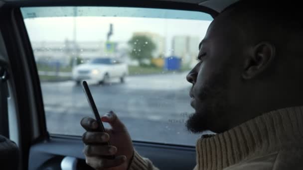 Retrato de hombre negro navegando teléfono inteligente está montando un coche en día lluvioso en la ciudad . — Vídeos de Stock