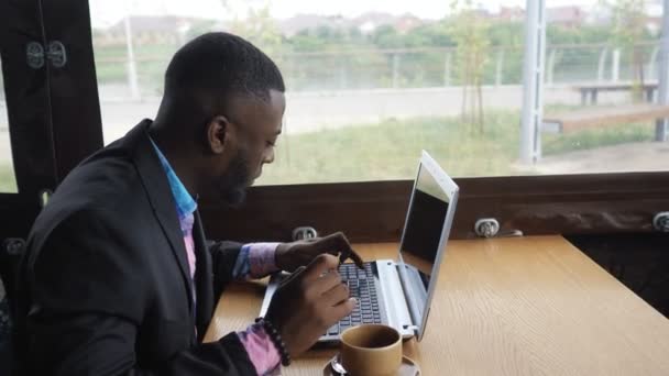 Black businessman is working typing a message on laptop sitting in summer cafe. — Stock Video