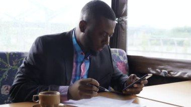 Afro Amerikalı işadamı akıllı telefon bakarak yaz çadır kafede documets dolduruyor.