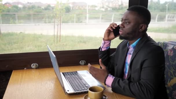 Fekete üzletember dolgozik laptop és felhívja smartphone ül kávézóban kávéval. — Stock videók