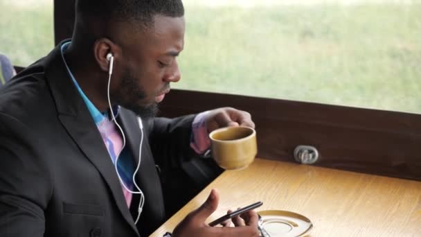 Uomo nero sta ascoltando musica in cuffia navigazione smartphone seduto in caffè . — Video Stock