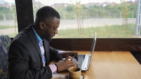 Uomo d'affari nero sta lavorando digitando un messaggio sul computer portatile seduto nel caffè estivo . — Foto Stock