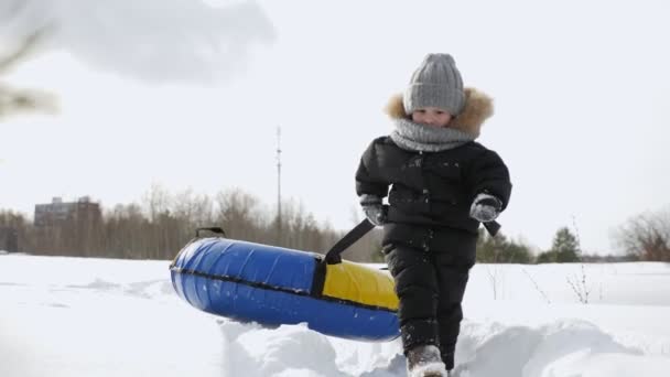 小さな男の子は冬の公園で雪の中で滑るためのチューブを引っ張って歩いています. — ストック動画