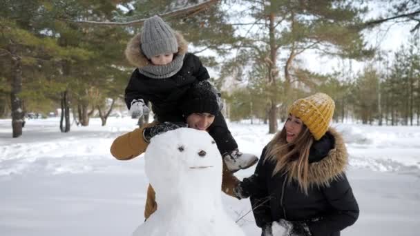Familia joven, mamá, hijo y papá están construyendo un muñeco de nieve en el parque de la ciudad de invierno . — Vídeos de Stock