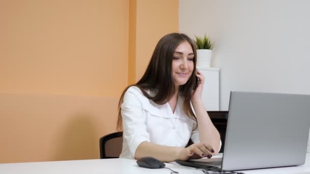 Lachende dame receptioniste praat op smartphone bij laptop — Stockvideo