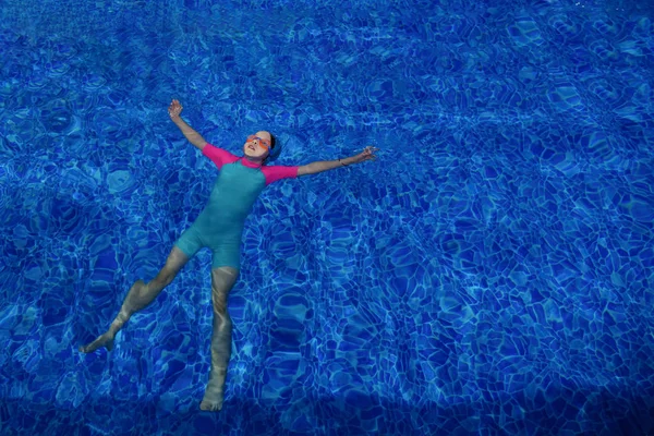 Enfant fille en lunettes, maillot de bain et casquette se détend sur le dos dans la piscine . — Photo