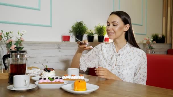 Smiling girl takes picture of cakes and tea at table in cafe — Stock Video