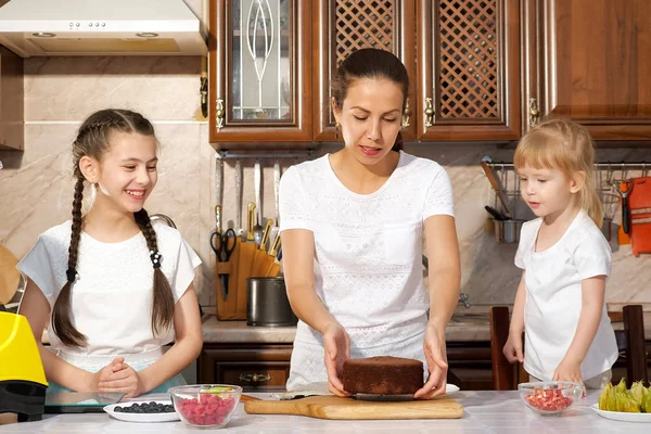 Mom is baking a cake with her two little daughters in the kitchen.