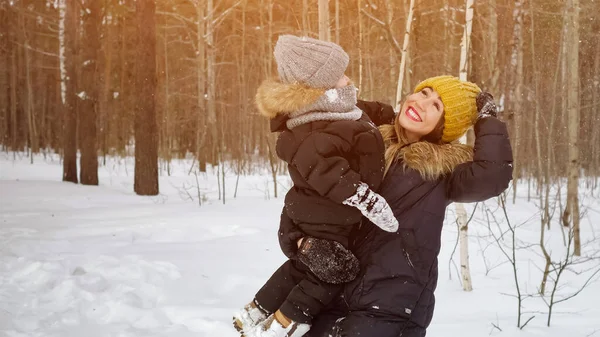 Jovem mãe com filho em suas mãos está olhando para a queda de neve na floresta de inverno . — Fotografia de Stock