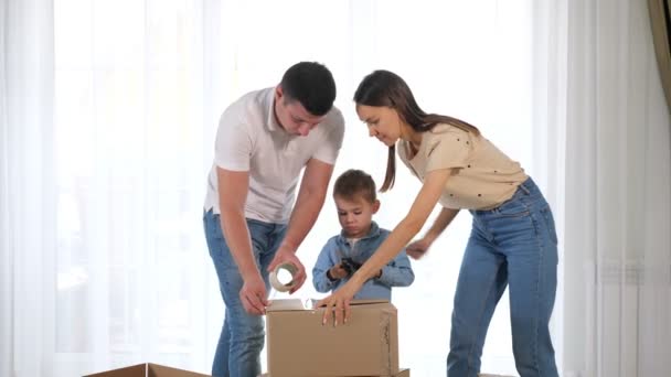 Joven pareja ayuda pequeño hijo paquete grande caja con cinta adhesiva — Vídeos de Stock
