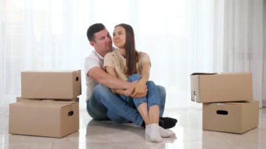 Kot pantolonlu bir erkek, yeni bir evde aydınlık bir zeminde oturup sarılıyor.