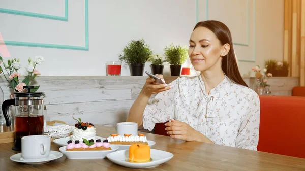 smiling girl takes picture of cakes and tea at table in cafe