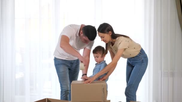 Mladý pár pomáhá malý syn balení velký box s lepicí páskou — Stock video