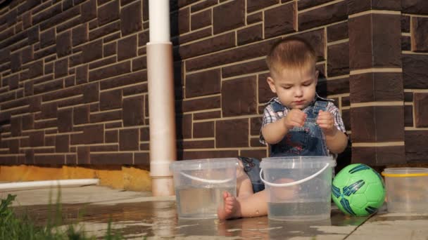 Мальчик с короткими светлыми волосами наливает воду из пластикового ведра — стоковое видео