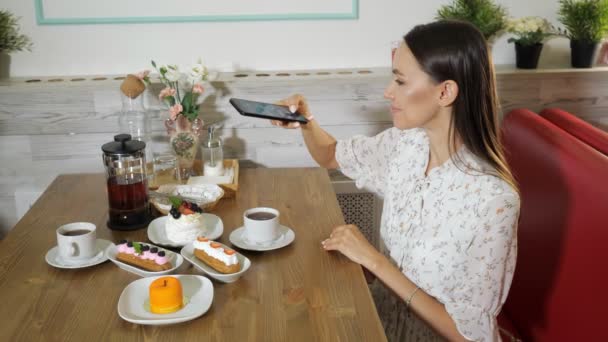 Chica sonriente toma fotos de pasteles y té en la mesa en la cafetería — Vídeos de Stock