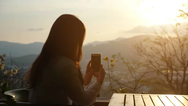 Young woman photographs sunset on the phone in a cafe on a hill