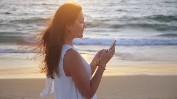 Flicka skriva på smartphone på havet stranden vid solnedgången — Stockvideo