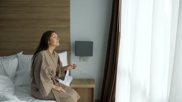 Heureuse dame en peignoir boit l'eau par la fenêtre de la chambre d'hôtel — Video