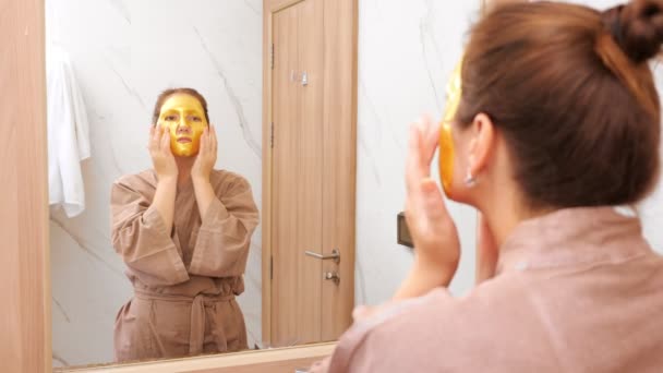 Vrouw met gouden masker doet gezicht massage in hotel spiegel — Stockvideo