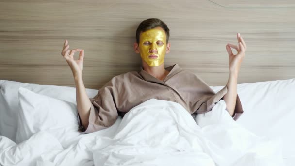 Мужчина в пальто с золотой маской для лица медитирует в постели в отеле — стоковое видео