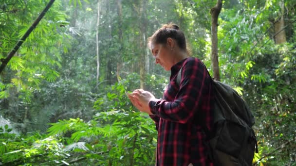 Señora sostiene el teléfono inteligente en la mano y mira a su alrededor en la selva tropical — Vídeos de Stock