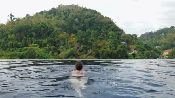 Lady swims to pool edge against picturesque hill with forest — Stock Video