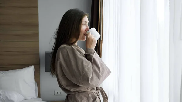 Fille en peignoir boit café près de la fenêtre dans la chambre d'hôtel — Photo