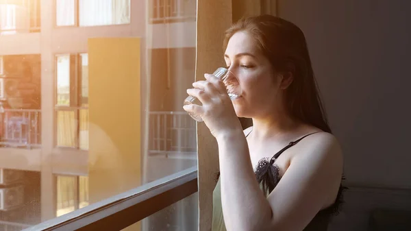 Счастливая дама пьет воду, глядя на окно в номере отеля — стоковое фото