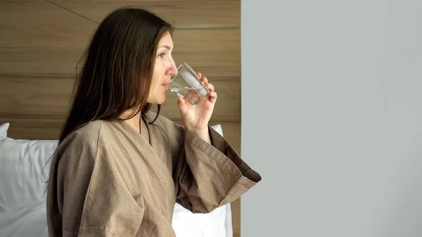 Lykkelig dame i badekåbe drikker vand ved hotelværelse vindue - Stock-foto