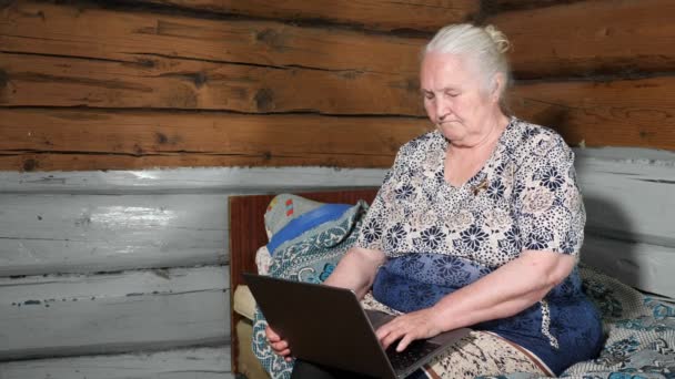 Anciana con el pelo gris sostiene un ordenador portátil en su regazo sentado en una cama — Vídeos de Stock