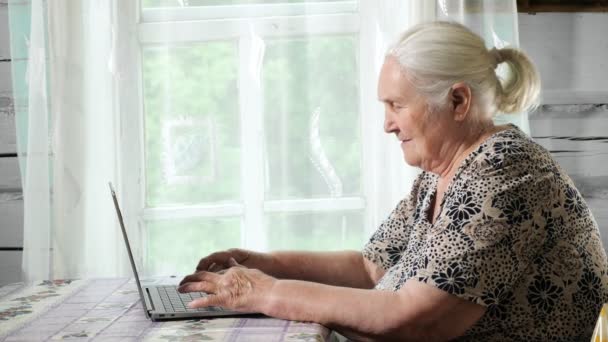 Pencerenin yanındaki masada oturan dizüstü bilgisayarda yazan yaşlı bir kadın. Yavaş çekim — Stok video