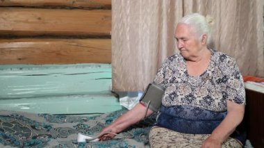 Yaşlı kadın tansiyon monitörüyle basıncı ölçüyor, yatakta oturuyor.