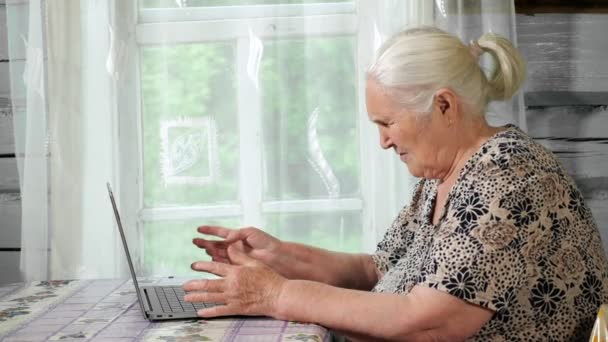 Mujer mayor escribiendo en un portátil sentado en una mesa junto a la ventana — Vídeos de Stock