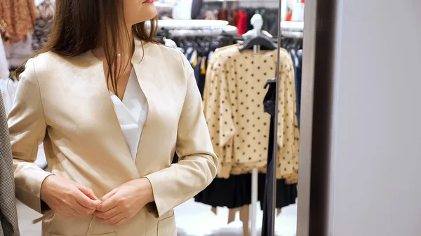 Mujer joven probándose una chaqueta crema con un cinturón delgado delante de un espejo en una tienda — Foto de Stock