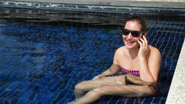 Bikinili kadın havuz suyunda oturarak akıllı telefondan konuşuyor. — Stok fotoğraf