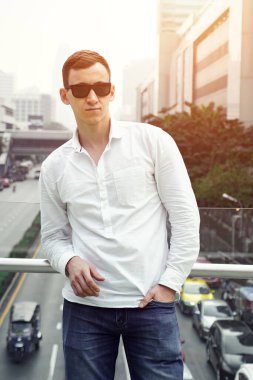 Güneş gözlüklü yakışıklı bir adam ve yol kenarındaki köprüde beyaz bir gömlek.